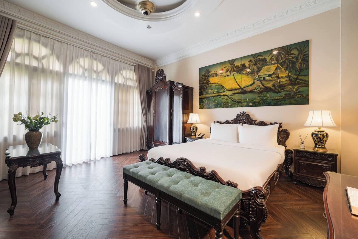 A large bed with ornate headboard, a bench, two bedside tables with lamps, a painting on the wall, and a large window with curtains.