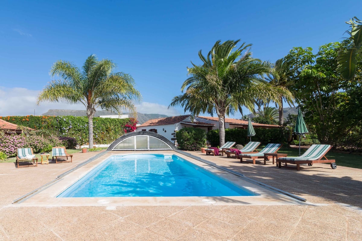 Una piscina con sillas de descanso y palmeras, rodeada de un jardín y un edificio con techo de tejas.