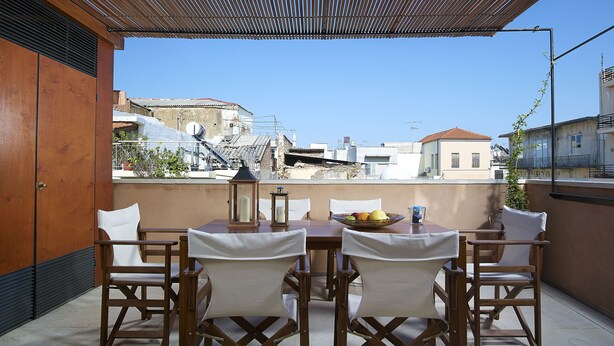 Eine Terrasse mit Holztisch und -stühlen, Blick auf Gebäude im Hintergrund und einer Pergola darüber.