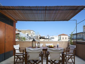 Una terraza con mesa y sillas de madera, vista de edificios al fondo y una pérgola arriba.
