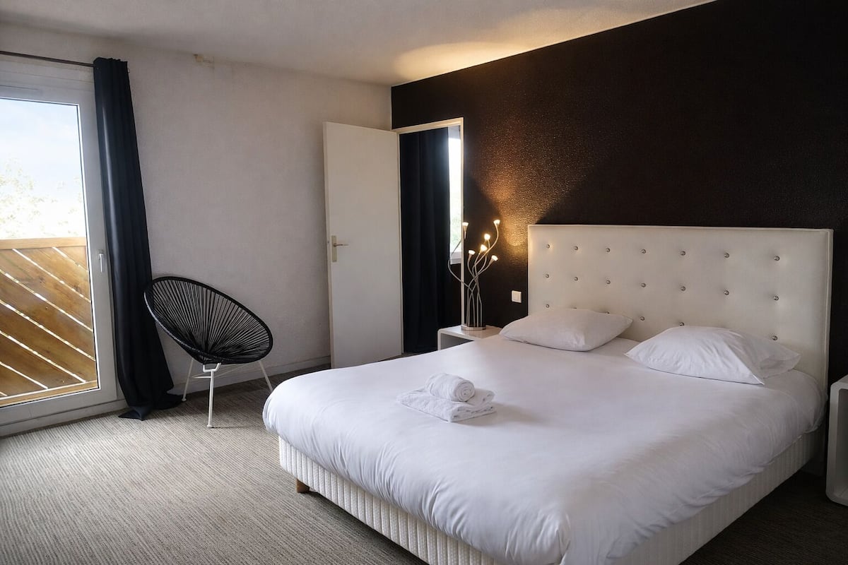 A hotel room with a large bed, a chair, a bedside table with a lamp, and a view through a sliding door.