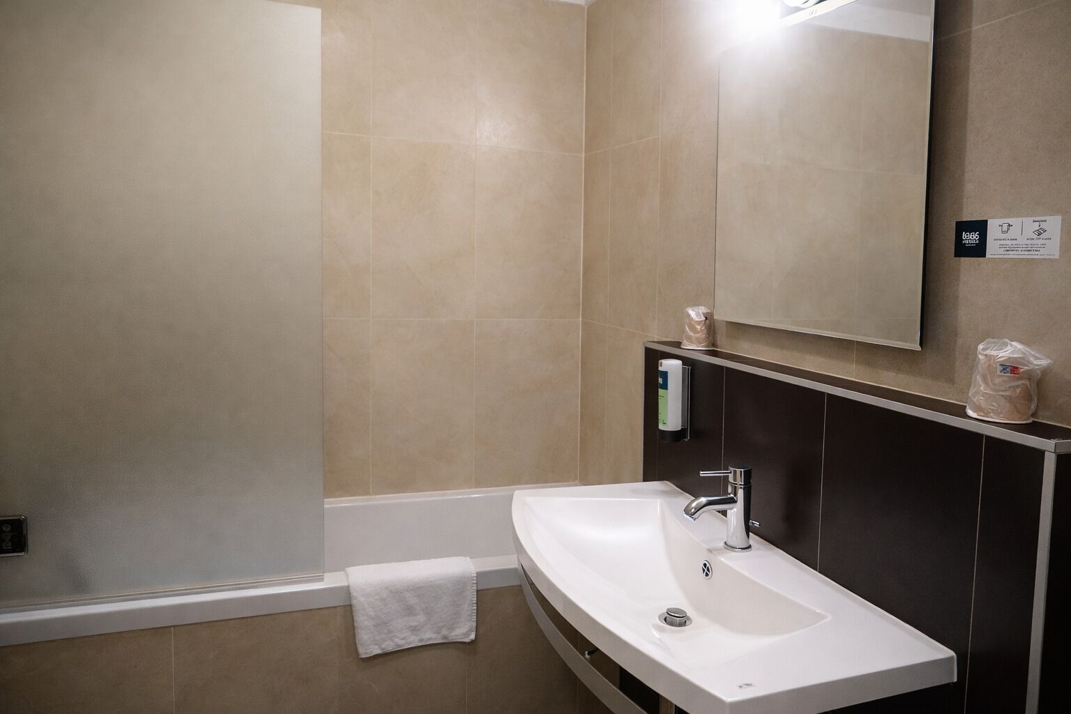 A modern bathroom with a white sink, dark wall paneling, a mirror, and a towel on the floor.
