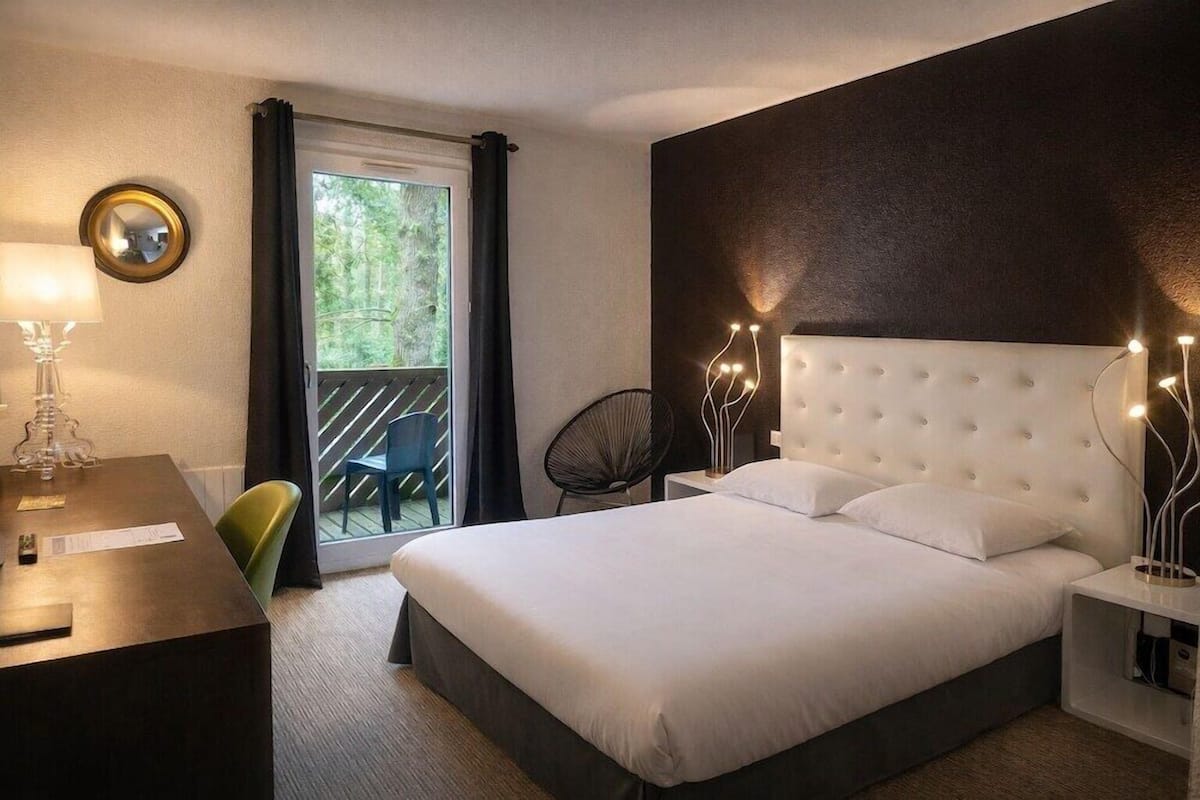 A hotel room with a bed, a desk, a chair, a balcony, and a view of greenery.