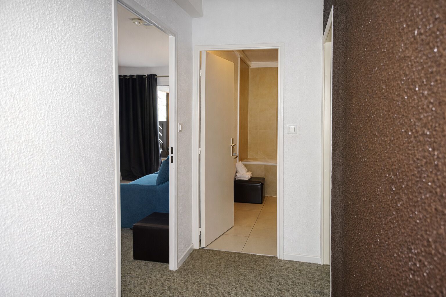 Un pasillo que conduce a un baño con ducha e inodoro. En la habitación contigua hay un sofá azul y un reposapiés negro.
