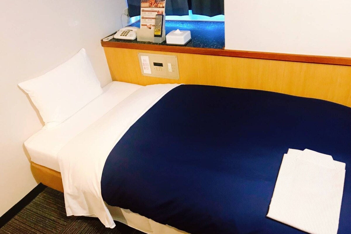 A hotel room with a single bed, a bedside table with a phone, a tissue box, and a small screen displaying a menu.