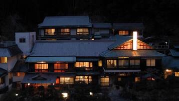 A traditional Japanese house with multiple levels, a prominent central chimney, and a well-lit exterior.