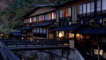 A traditional Japanese ryokan with multiple connected buildings, a wooden bridge, and a mountainous backdrop.