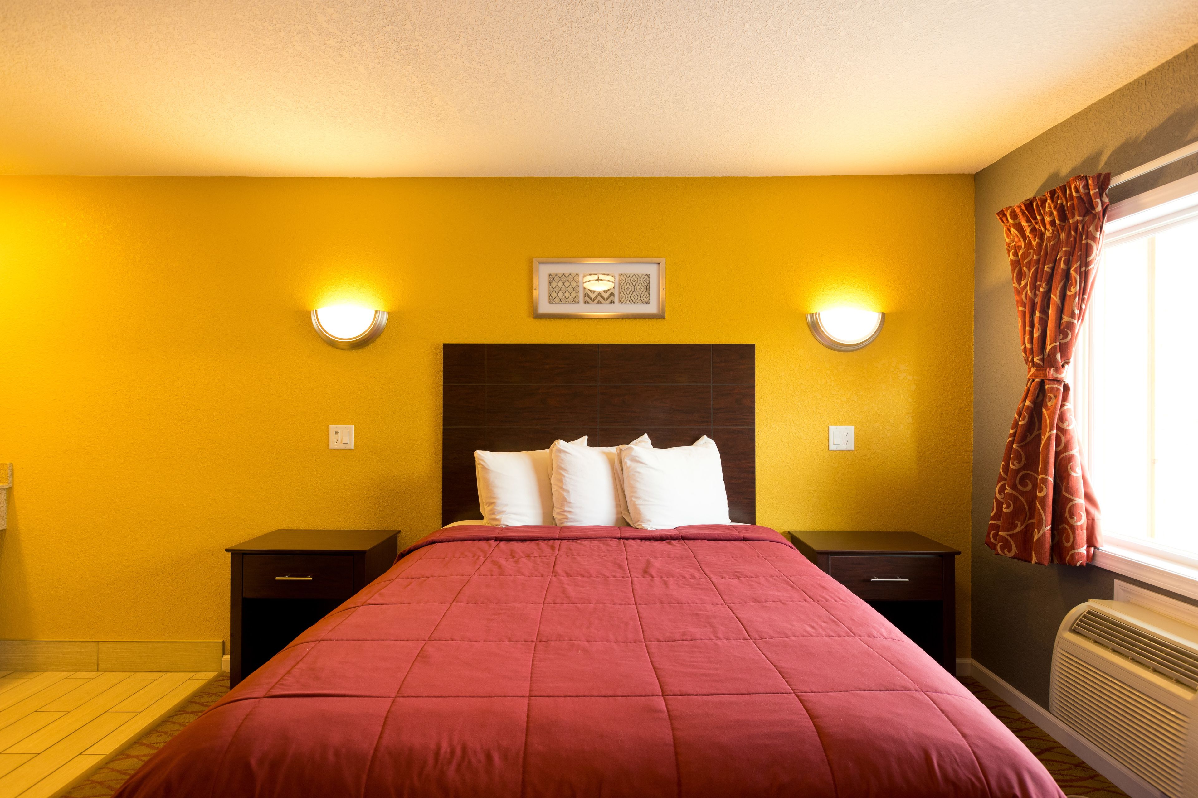 Une chambre d’hôtel comprenant un lit, deux tables de chevet, une petite table, un luminaire fixé au mur, une fenêtre avec des rideaux et un appareil de climatisation fixé au mur.