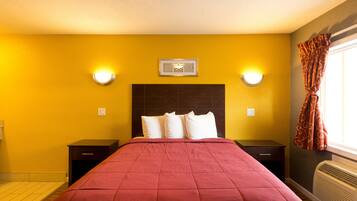 Une chambre d’hôtel comprenant un lit, deux tables de chevet, une petite table, un luminaire fixé au mur, une fenêtre avec des rideaux et un appareil de climatisation fixé au mur.