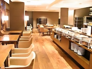Un café moderne avec des tables et des chaises en bois, un comptoir de buffet où sont disposés des récipients contenant de la nourriture, et un intérieur lumineux.