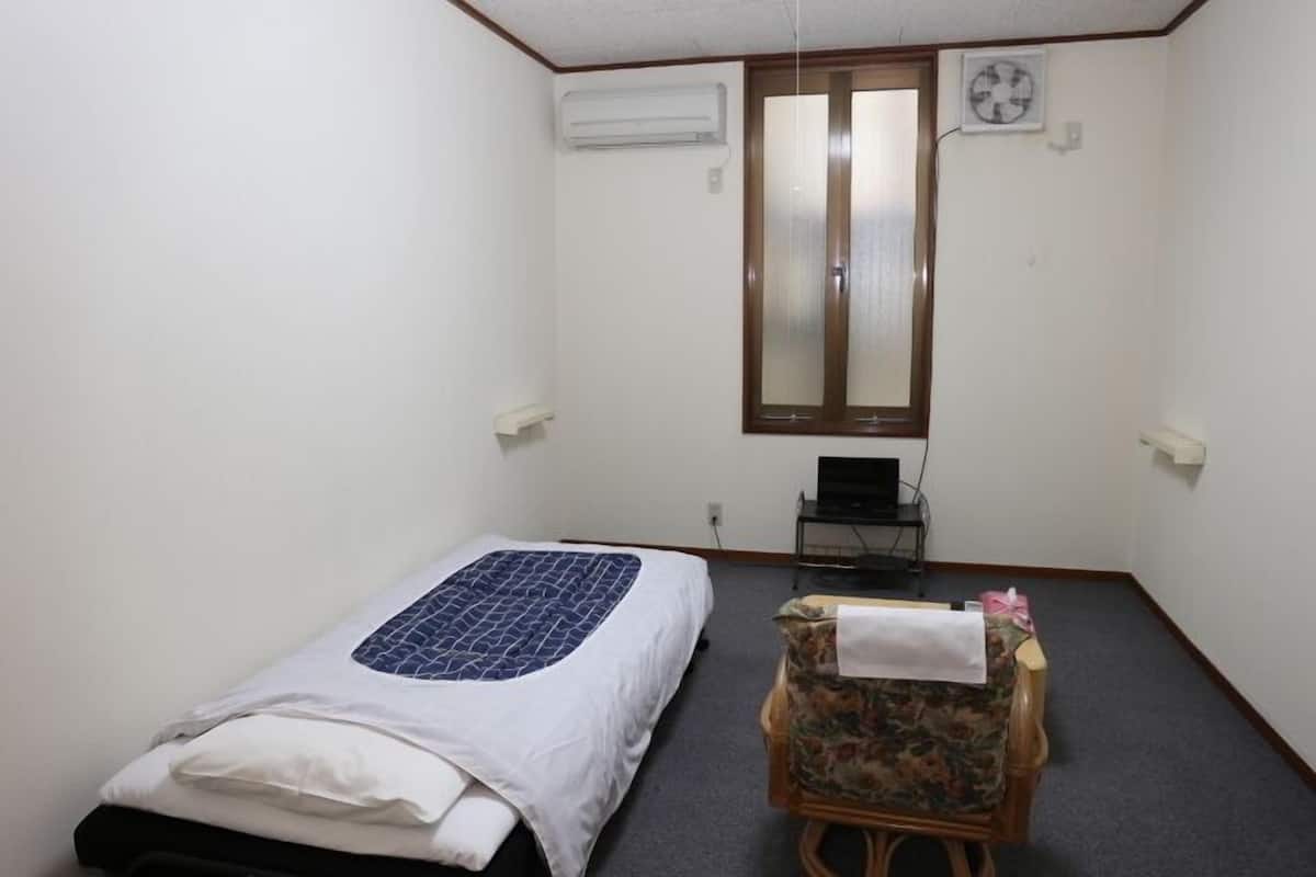 A small, simple room with a bed, a chair, an air conditioning unit, a window, and a desk with a computer.