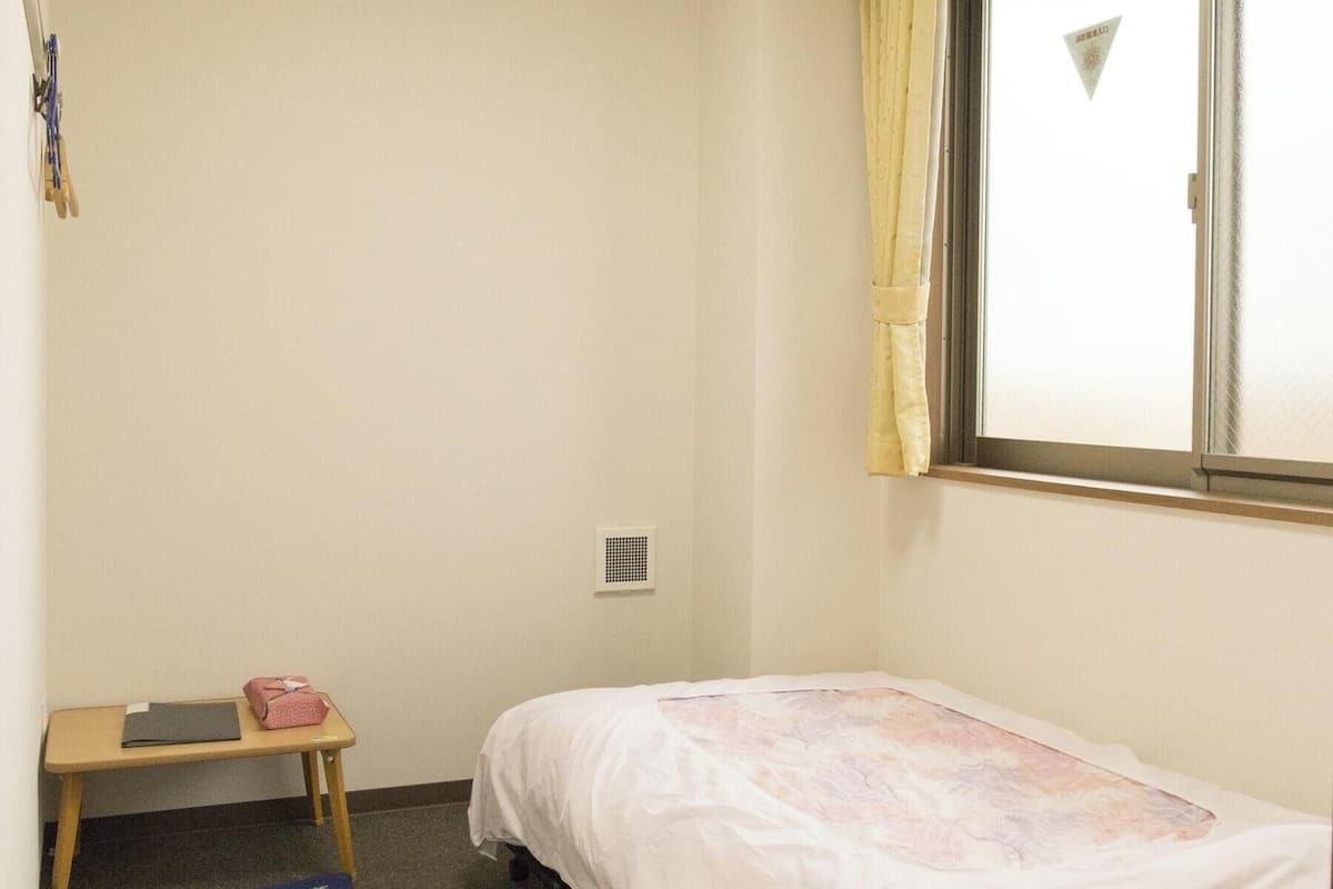 A small, sparsely furnished room with a single bed, a window with curtains, a small table with items on it, and a ceiling light.