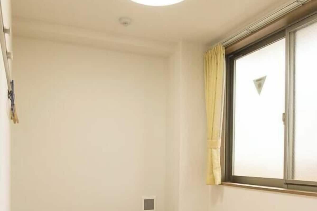 A small, sparsely furnished room with a single bed, a small table, a window with curtains, and a ceiling light.