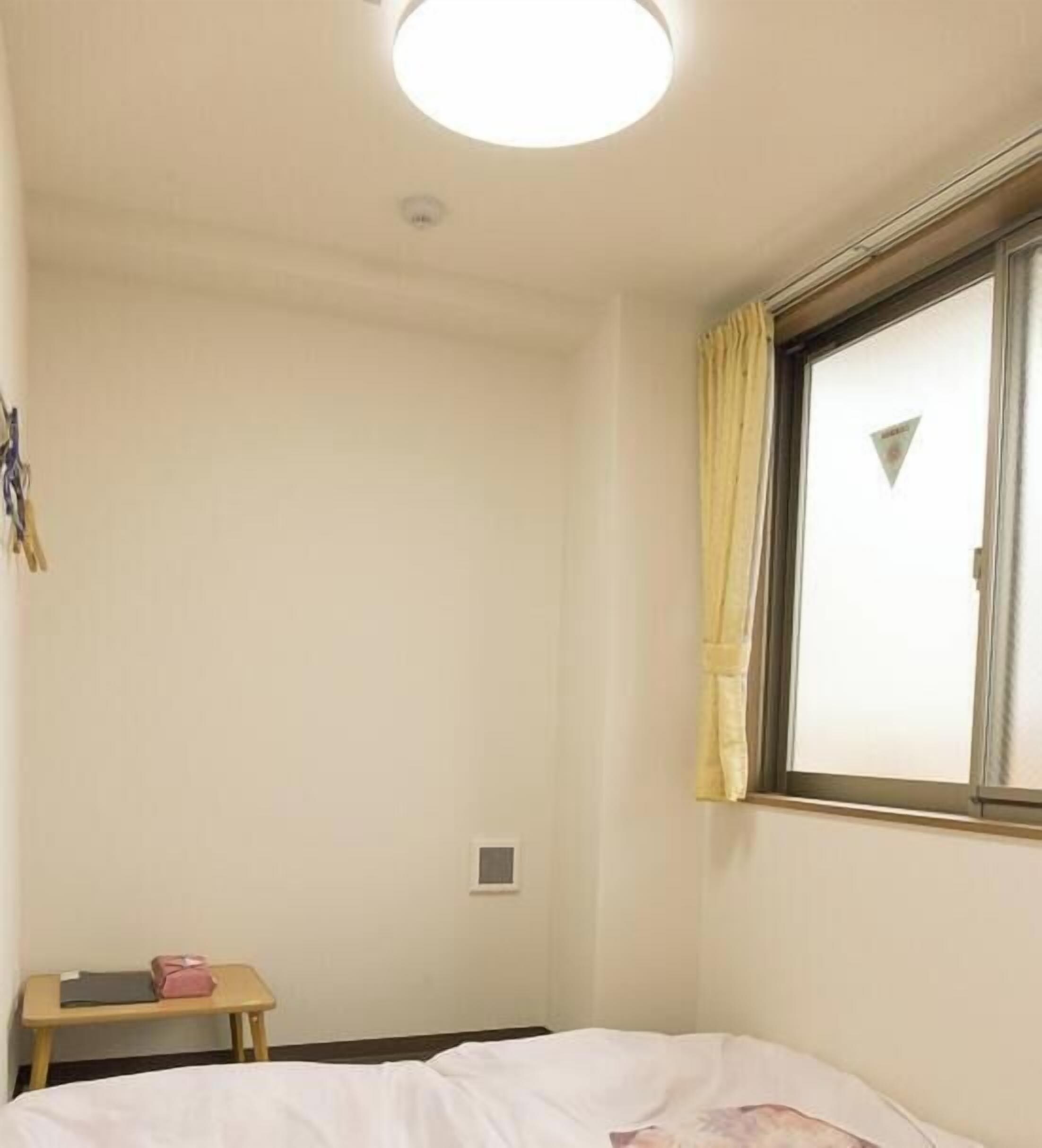 Een kleine, eenvoudige kamer met een bed, een klein tafeltje, een raam met gordijnen en een plafondlamp.