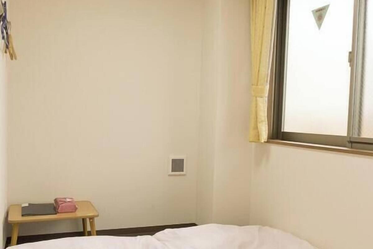 A small, simple room with a bed, a small table, a window with curtains, and a ceiling light.