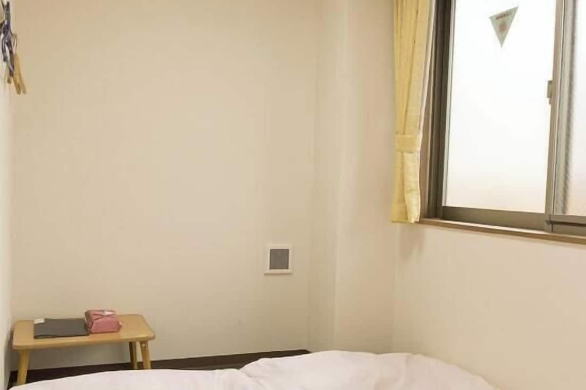 A small, simple room with a bed, a small table, a window with curtains, and a ceiling light.