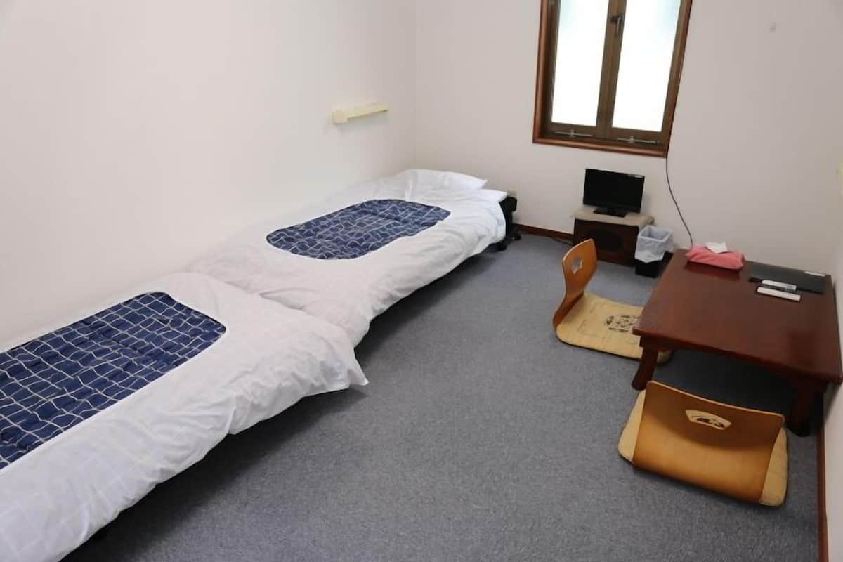 A small, single-room accommodation with two single beds, a desk, a chair, a TV, and a window.