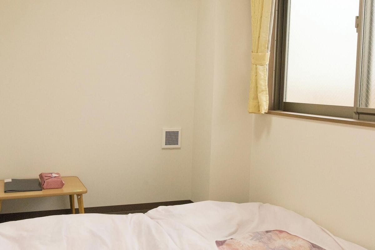 A room with a bed, a small table with items on it, a window with curtains, and a ceiling light.