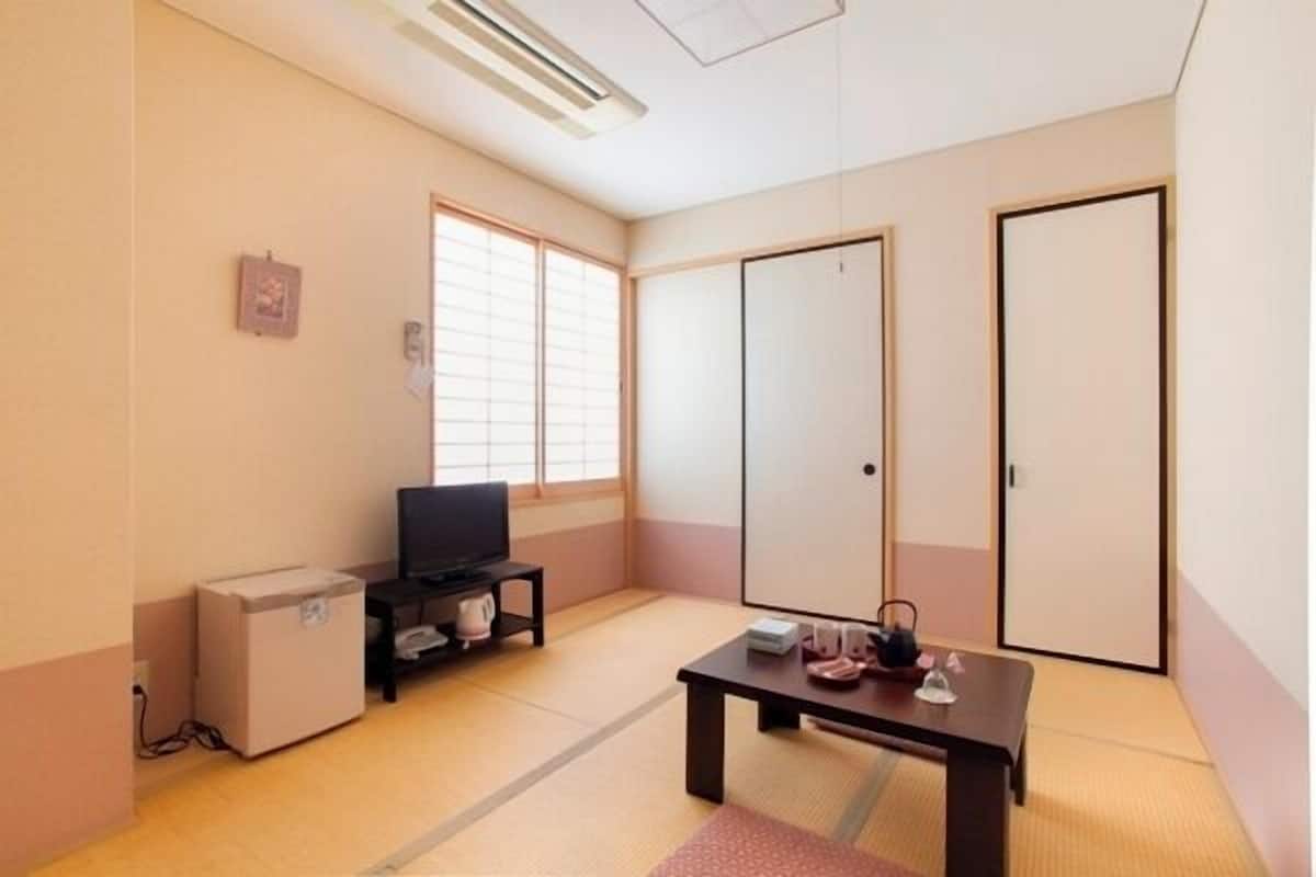 Une pièce avec un sol recouvert de tatamis, un téléviseur posé sur un meuble, une petite armoire, une table sur laquelle se trouvent divers objets et des portes coulissantes.