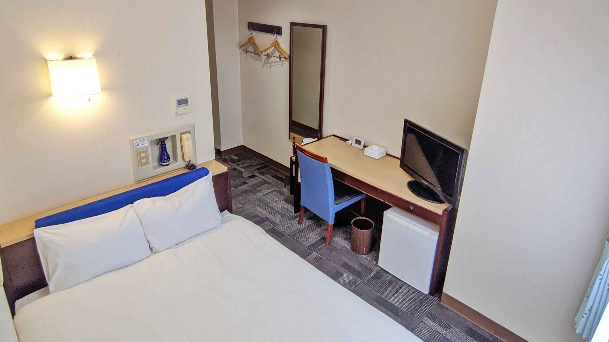 Quarto de hotel com cama, escrivaninha, cadeira, televisão, espelho e um pequeno cesto de lixo.