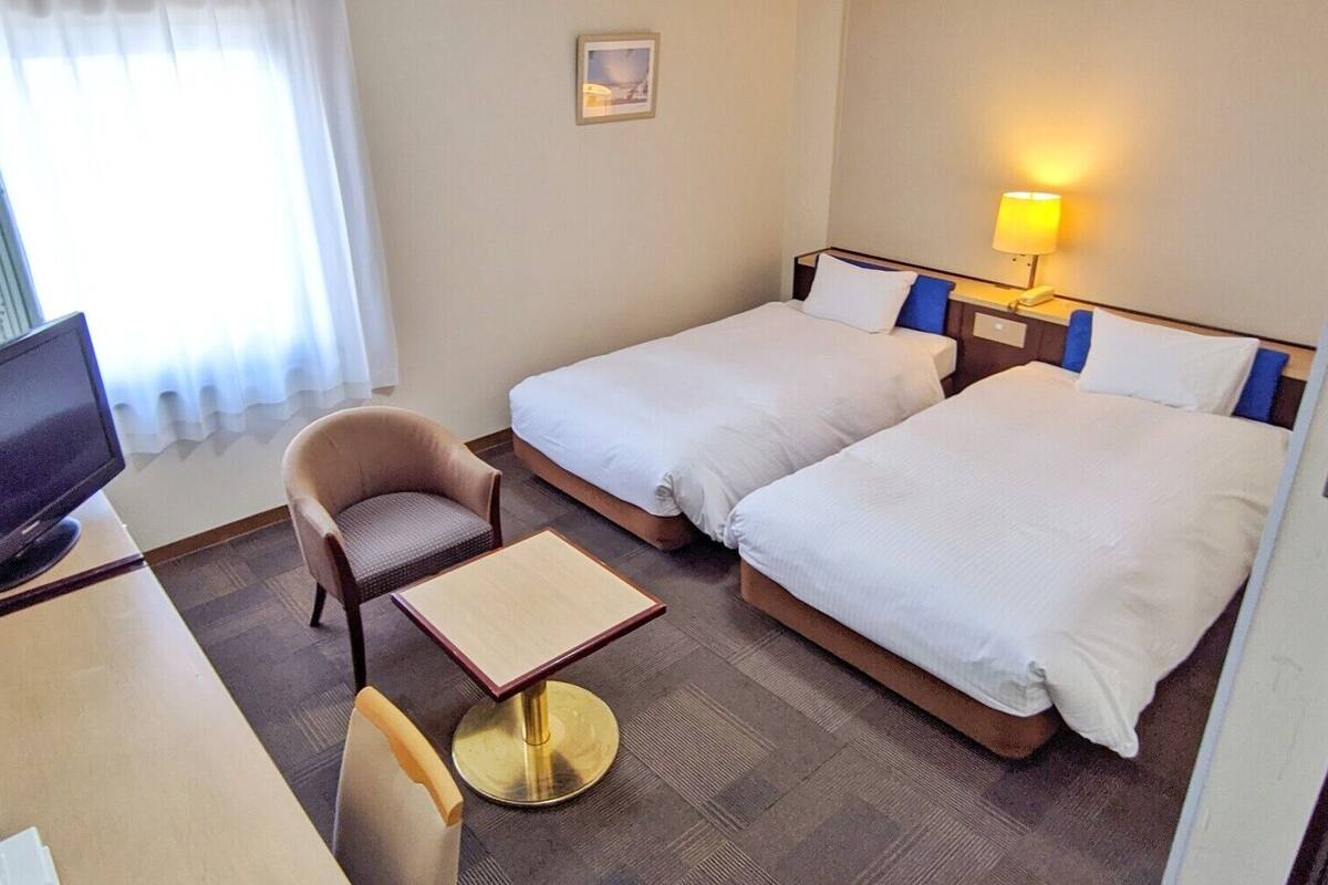 A hotel room with two beds, a TV, a chair, a small table, and a lamp.