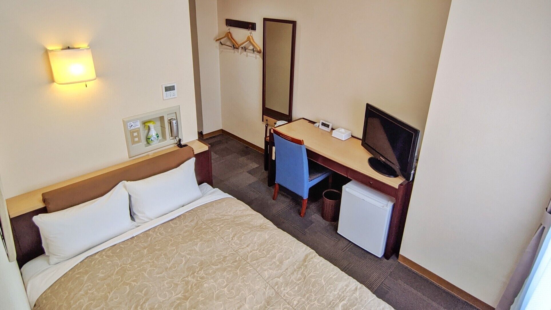 Quarto de hotel com cama, escrivaninha, cadeira, televisão e um pequeno frigorífico.
