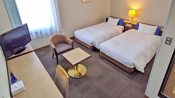 A hotel room with two beds, a desk with a television, a chair, and a small table. - Kadoma Public (Kadoma)