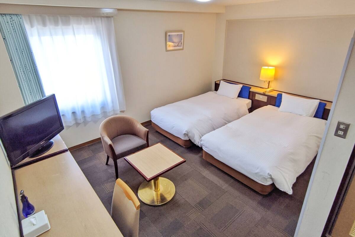 A hotel room with two beds, a desk with a television, a chair, a small table, and a lamp.
