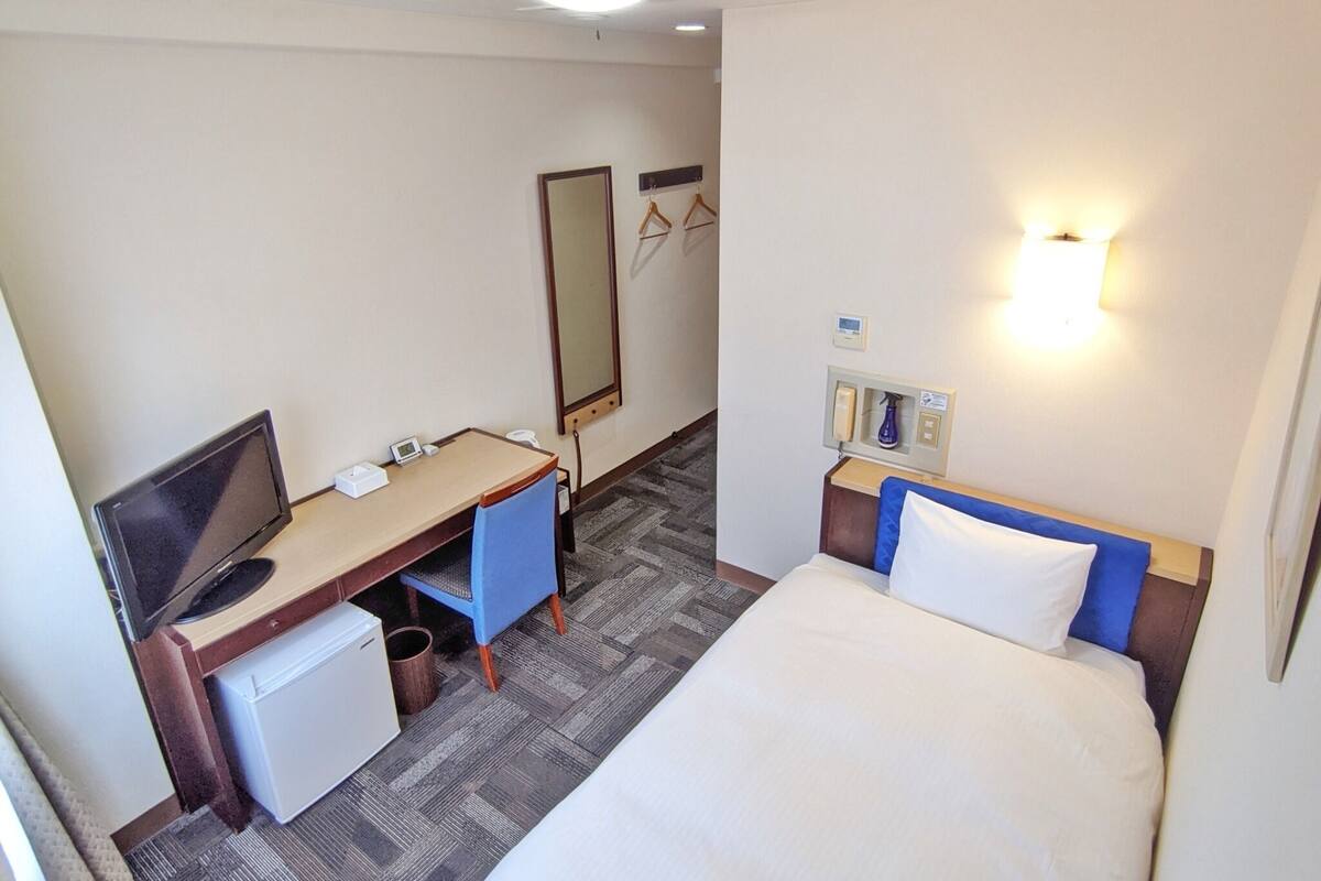 A hotel room with a bed, a desk with a computer, a chair, a small fridge, a mirror, and a wall-mounted light.