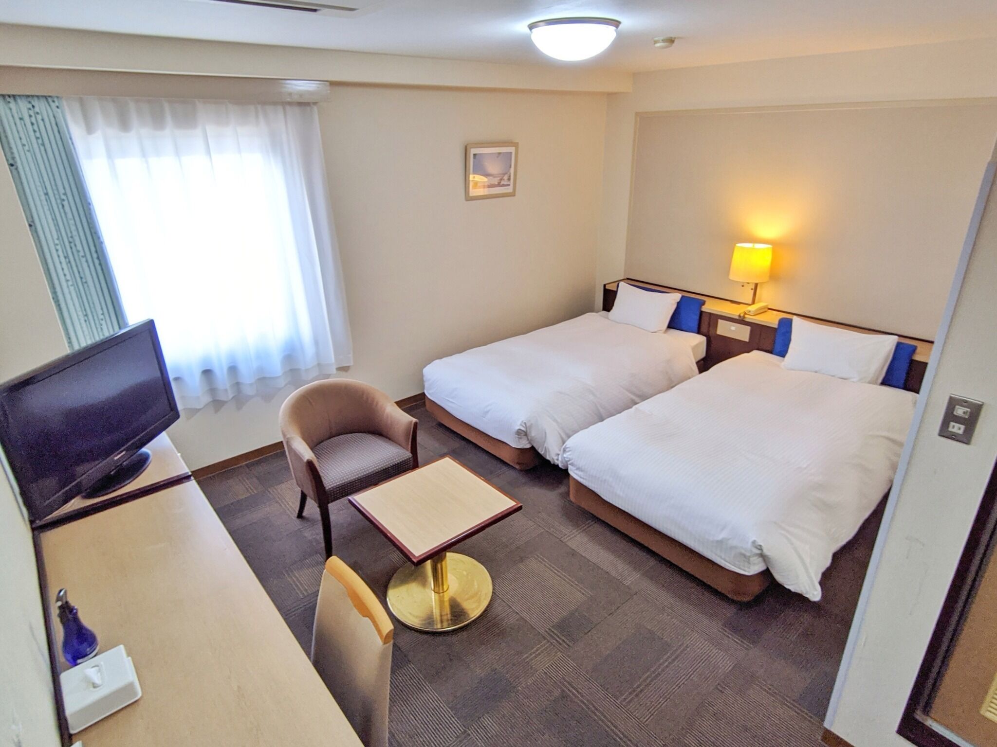 Quarto de hotel com duas camas, uma mesa de trabalho com televisão, uma cadeira, uma mesinha e um abajur.