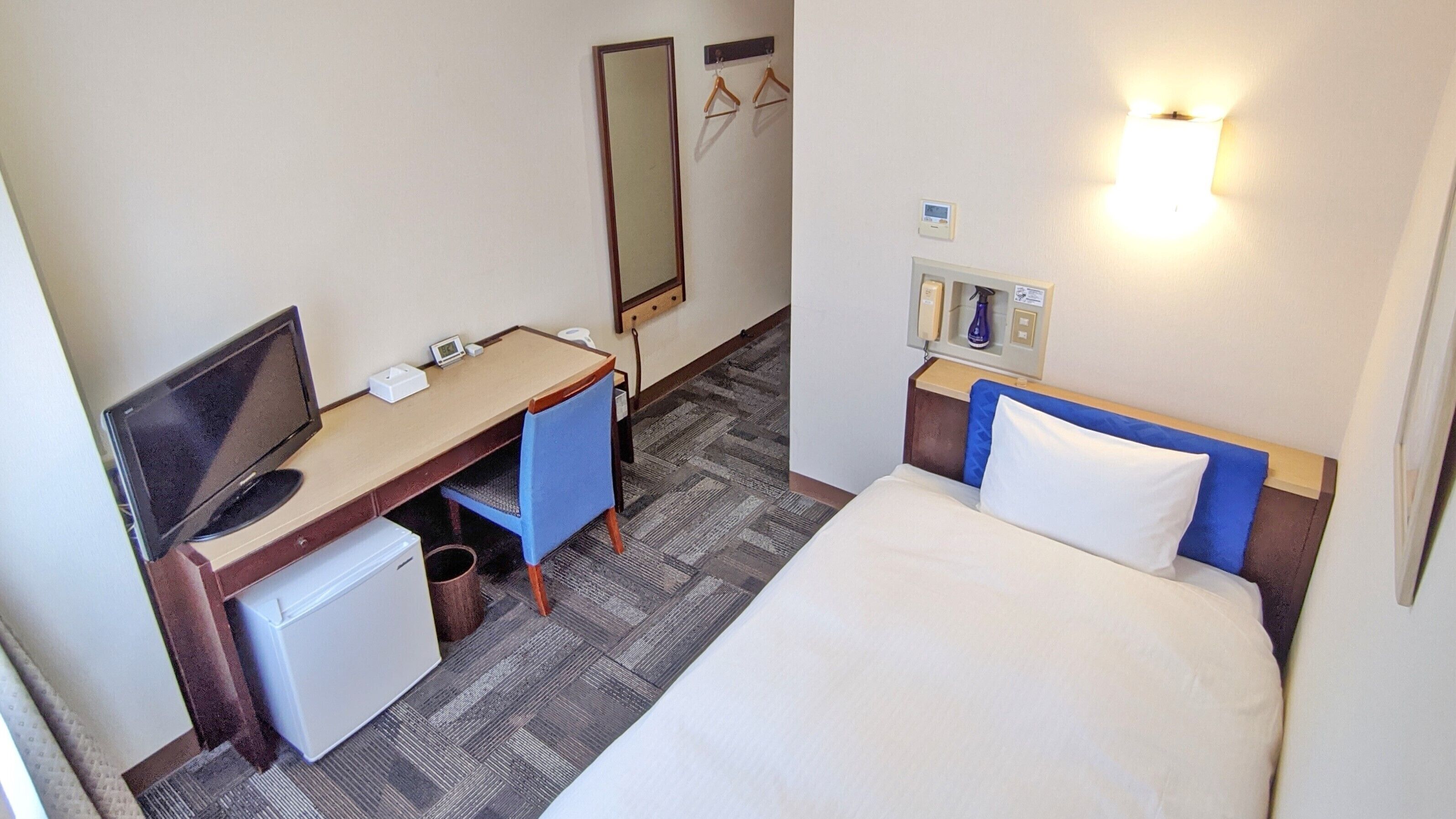 A hotel room with a bed, a desk with a computer, a small fridge, a mirror, and a wall-mounted light fixture.