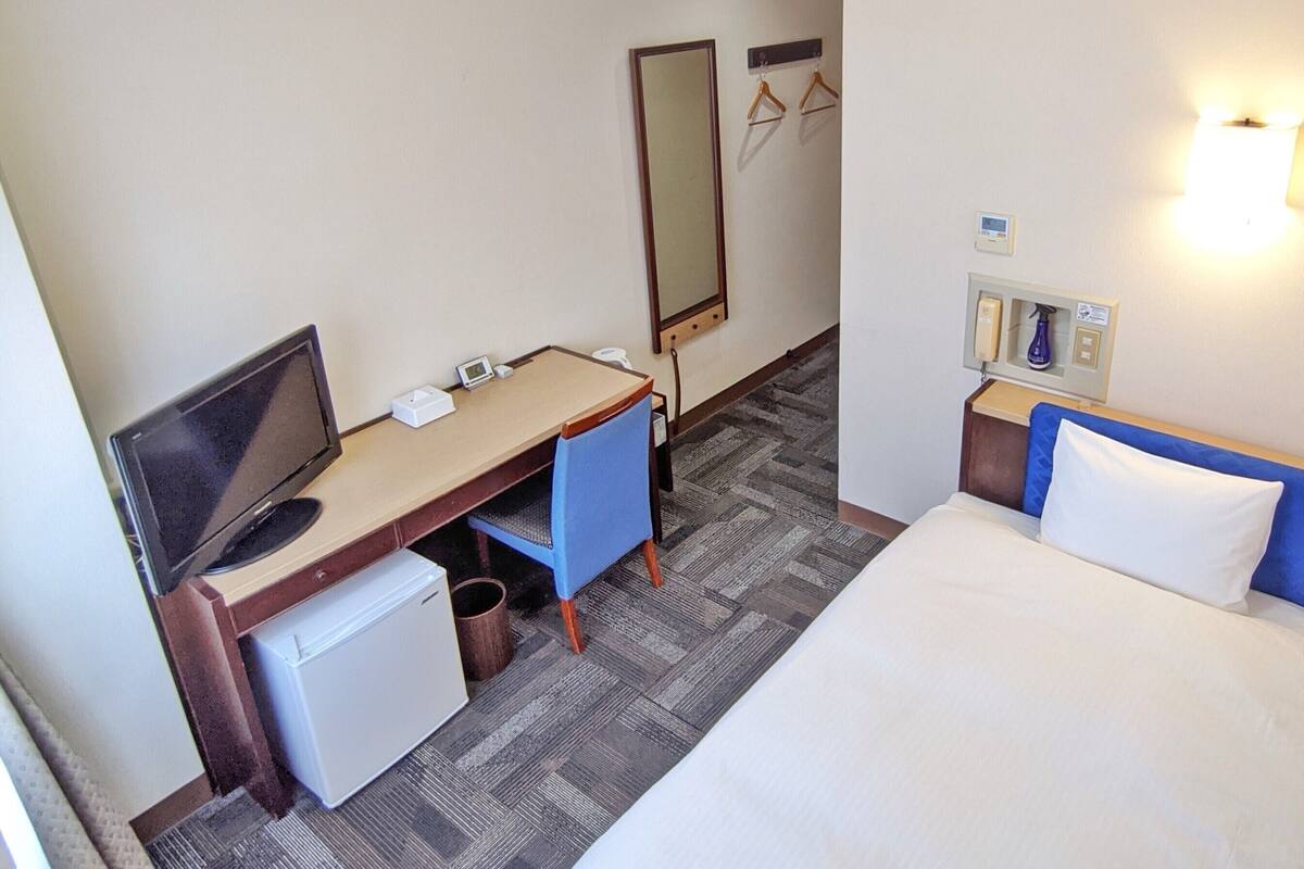 A hotel room with a bed, a desk with a computer, a small fridge, a mirror, and a wall-mounted light fixture.
