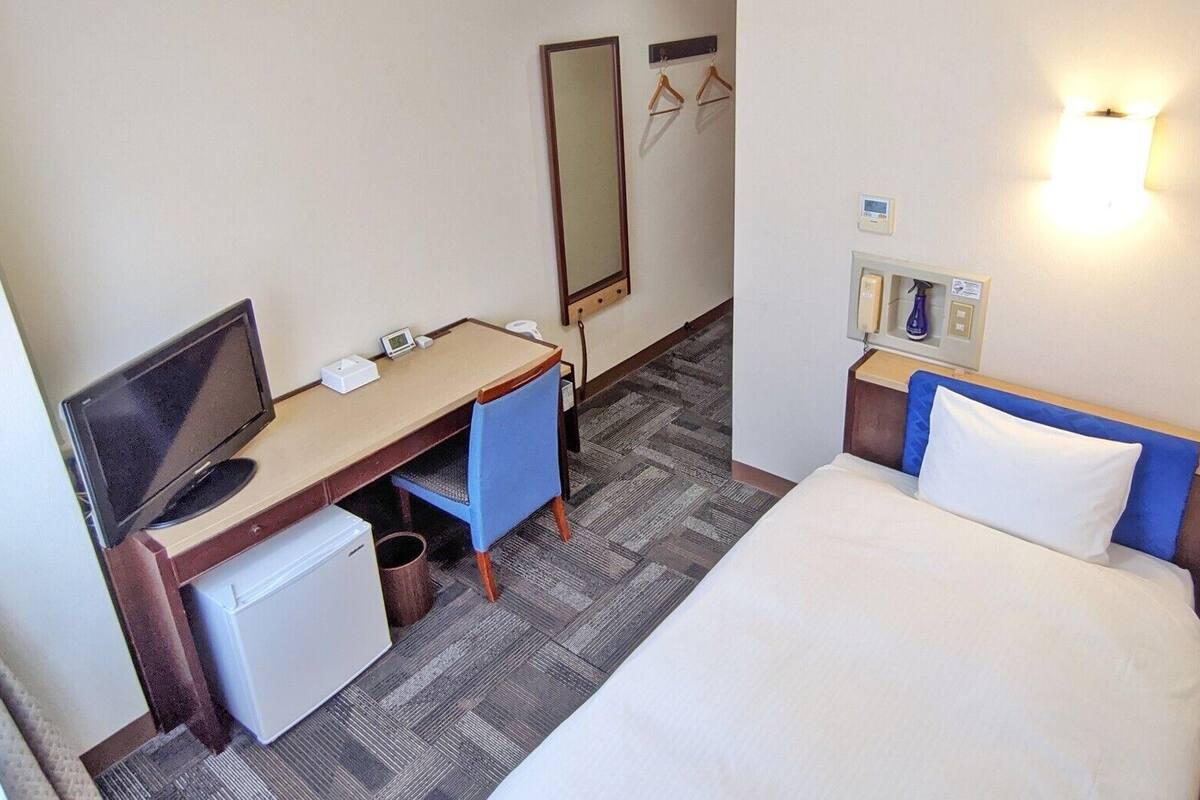 A hotel room with a bed, desk, chair, and a small refrigerator.