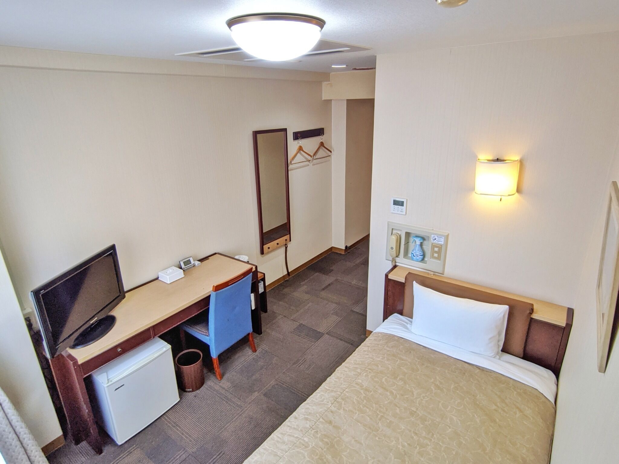 Quarto de hotel com cama, escrivaninha, cadeira, televisão e pequeno frigorífico.