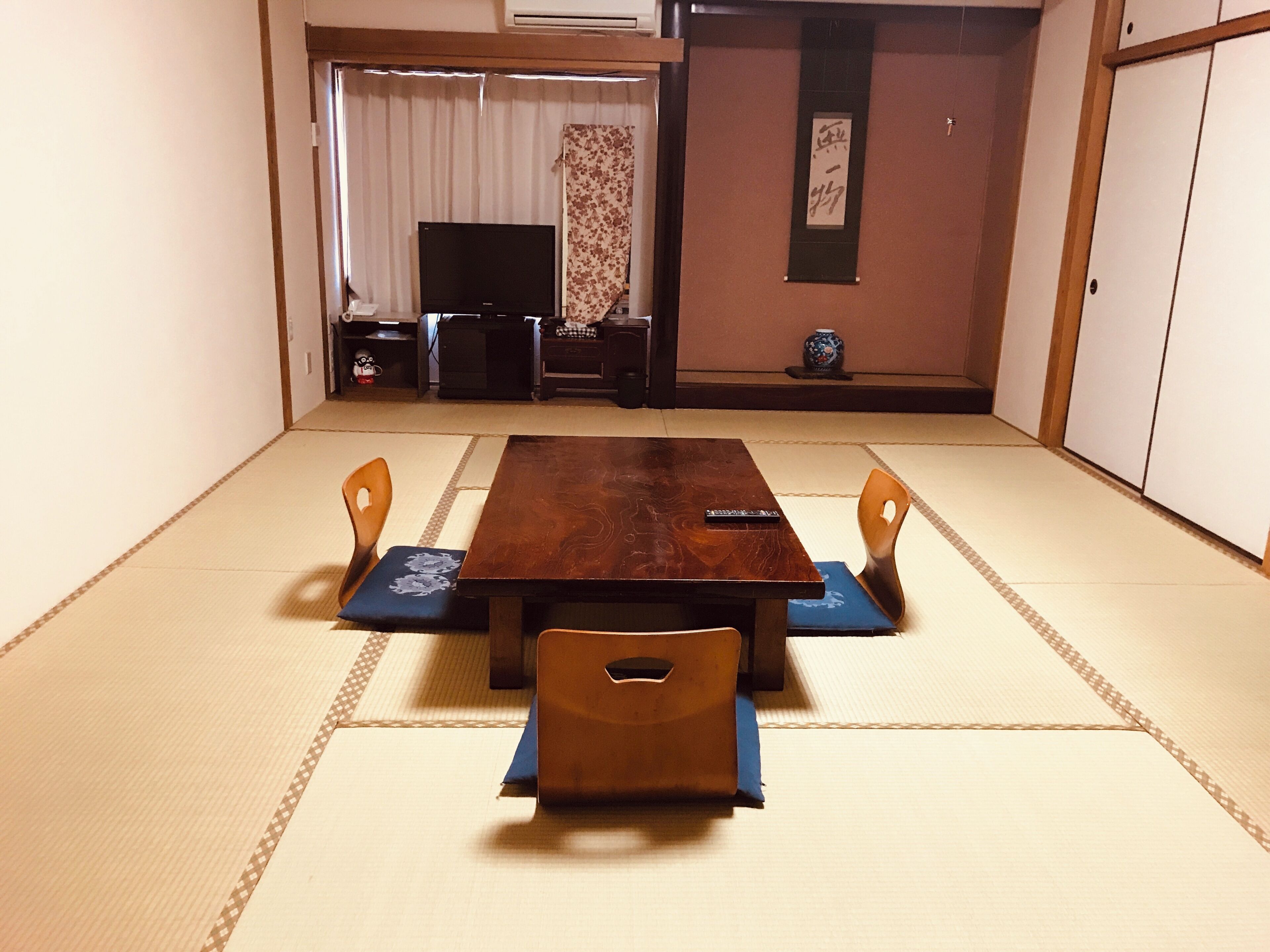 A tatami mat room with a wooden table and four chairs, a television, and a small decorative vase.