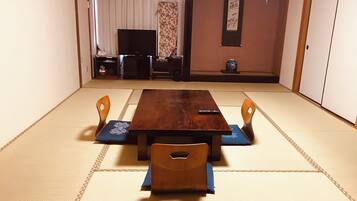 A tatami mat room with a wooden table and four chairs, a television, and a small decorative vase.