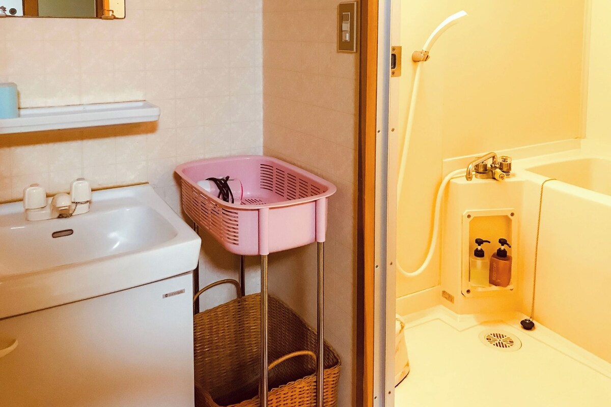 Un baño con un lavamanos blanco, una cesta rosa, una cesta de mimbre y una bañera con cabezal de ducha.
