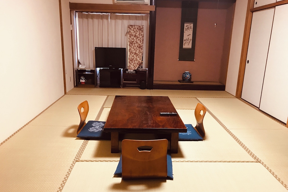 A tatami mat room with a wooden table and four chairs, a television, and a small decorative vase.