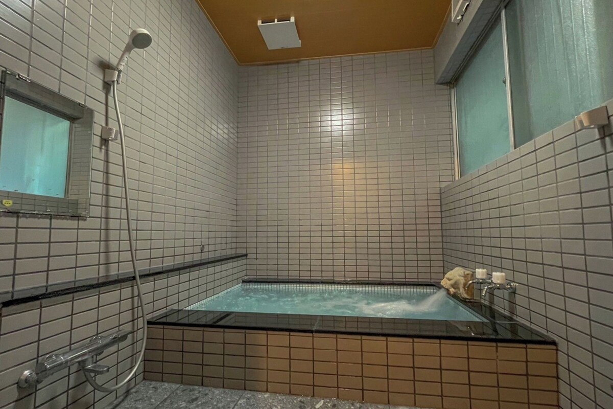 A bathroom with a glass-enclosed bathtub, a handheld showerhead, and a window with a curtain.