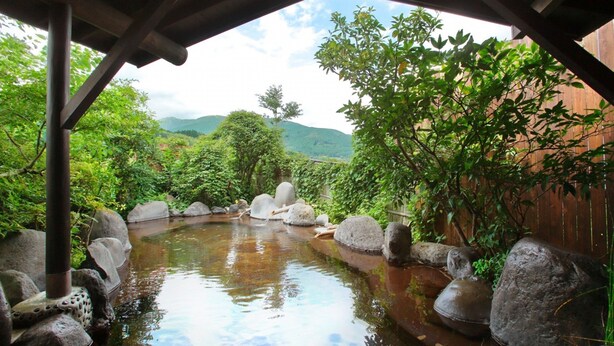 Un onsen japonés tradicional con estructura de madera, una piscina natural rodeada de rocas y abundante vegetación al fondo.