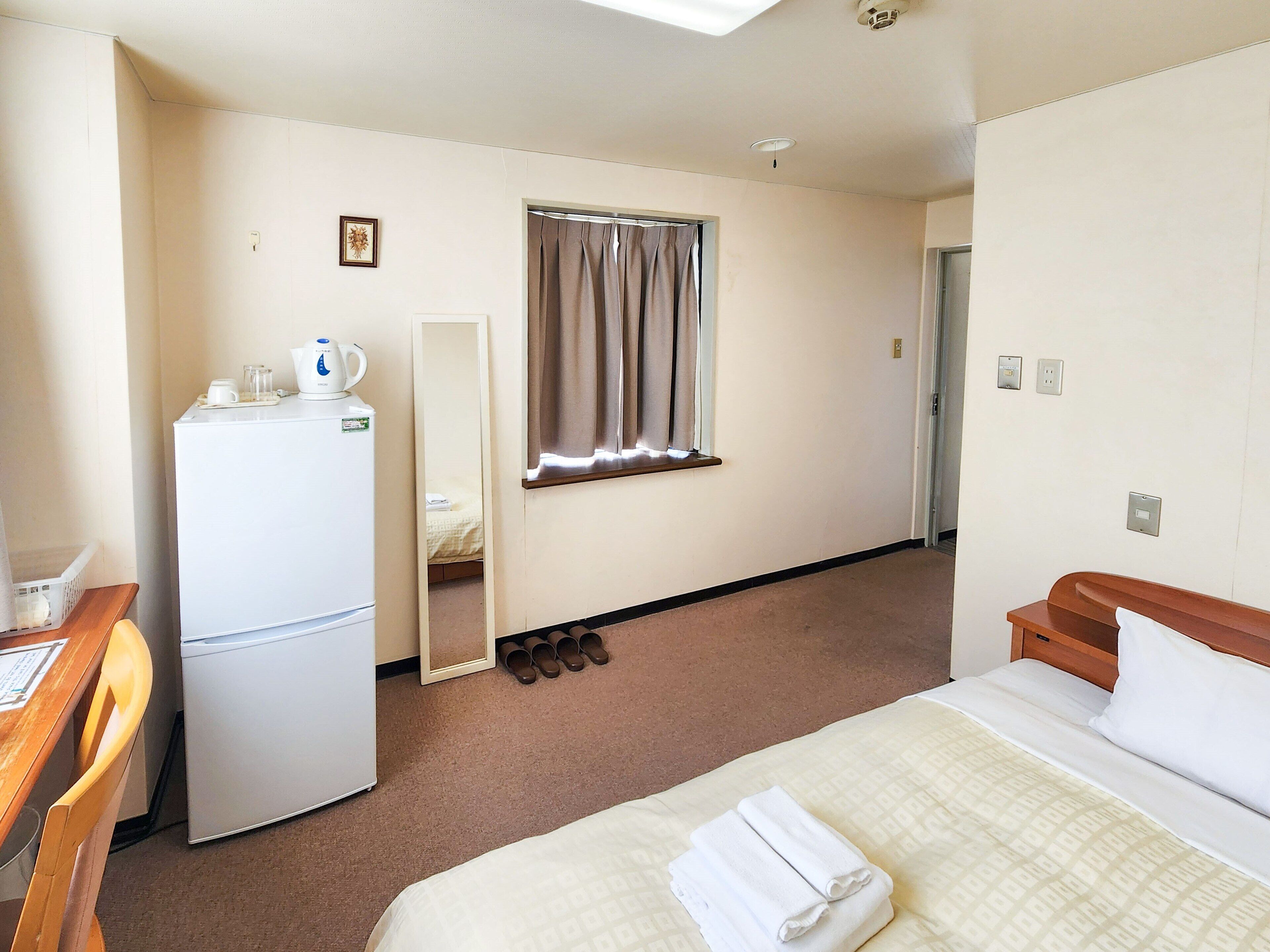 A hotel room with a bed, a desk with a chair, a refrigerator, a mirror, a window with curtains, and a door.