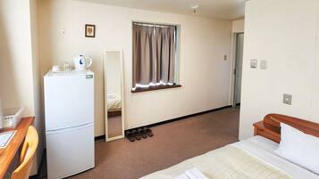 A hotel room with a bed, a desk with a chair, a refrigerator, a mirror, a window with curtains, and a door.