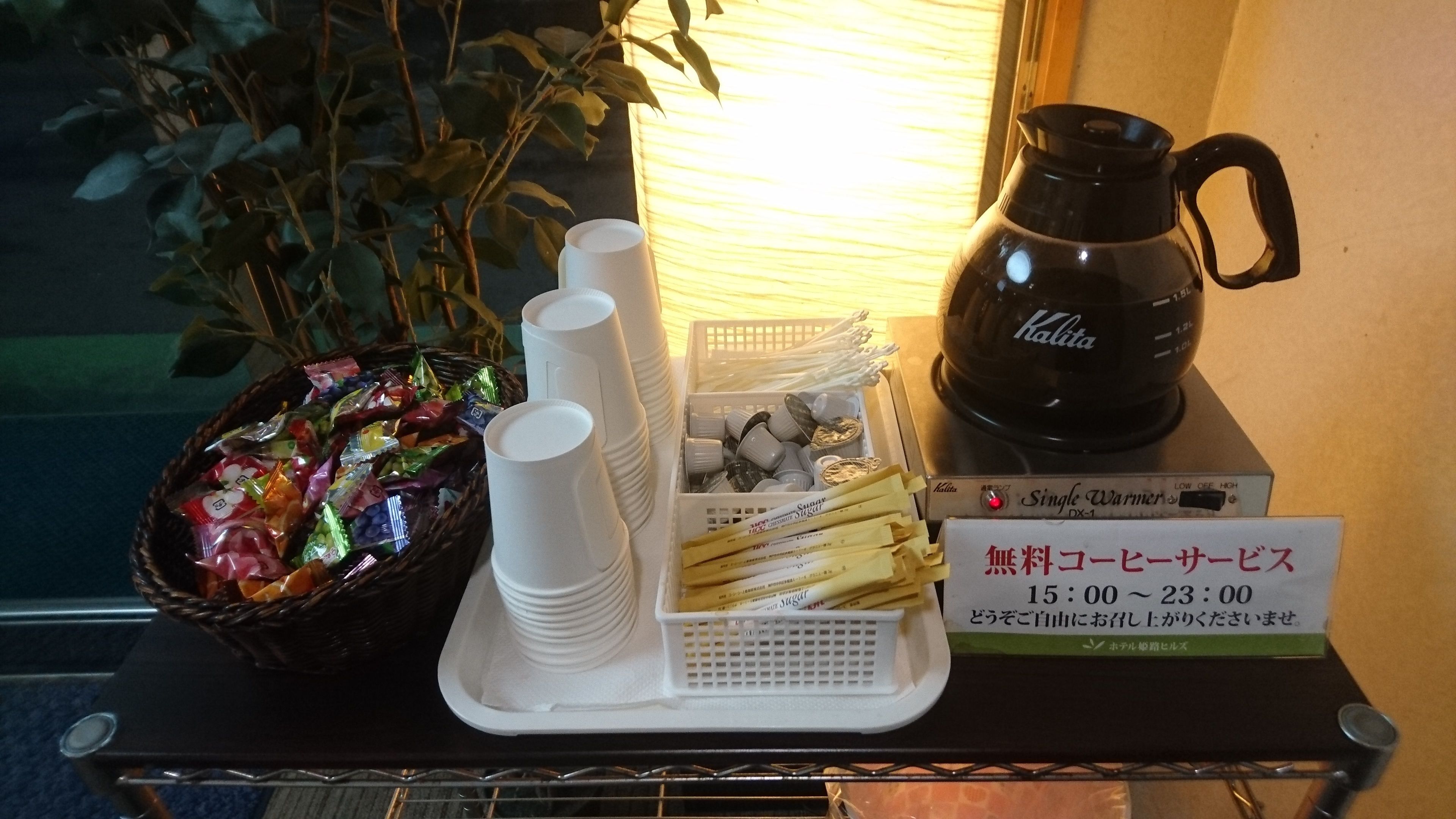 A tray with disposable cups, a basket of snacks, a coffee maker, and a sign indicating a free coffee service from 15:00 to 23:00.