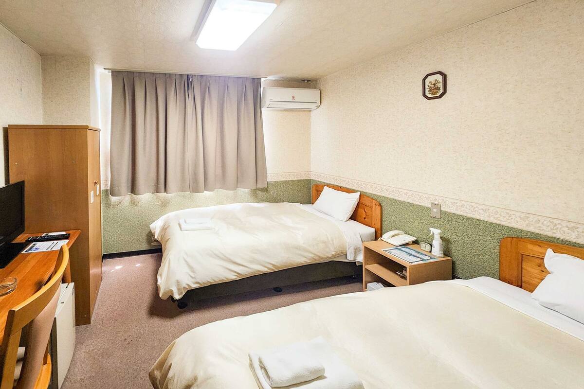 Habitación de hotel con dos camas, un escritorio, una silla, un televisor, aire acondicionado y una ventana con cortinas.