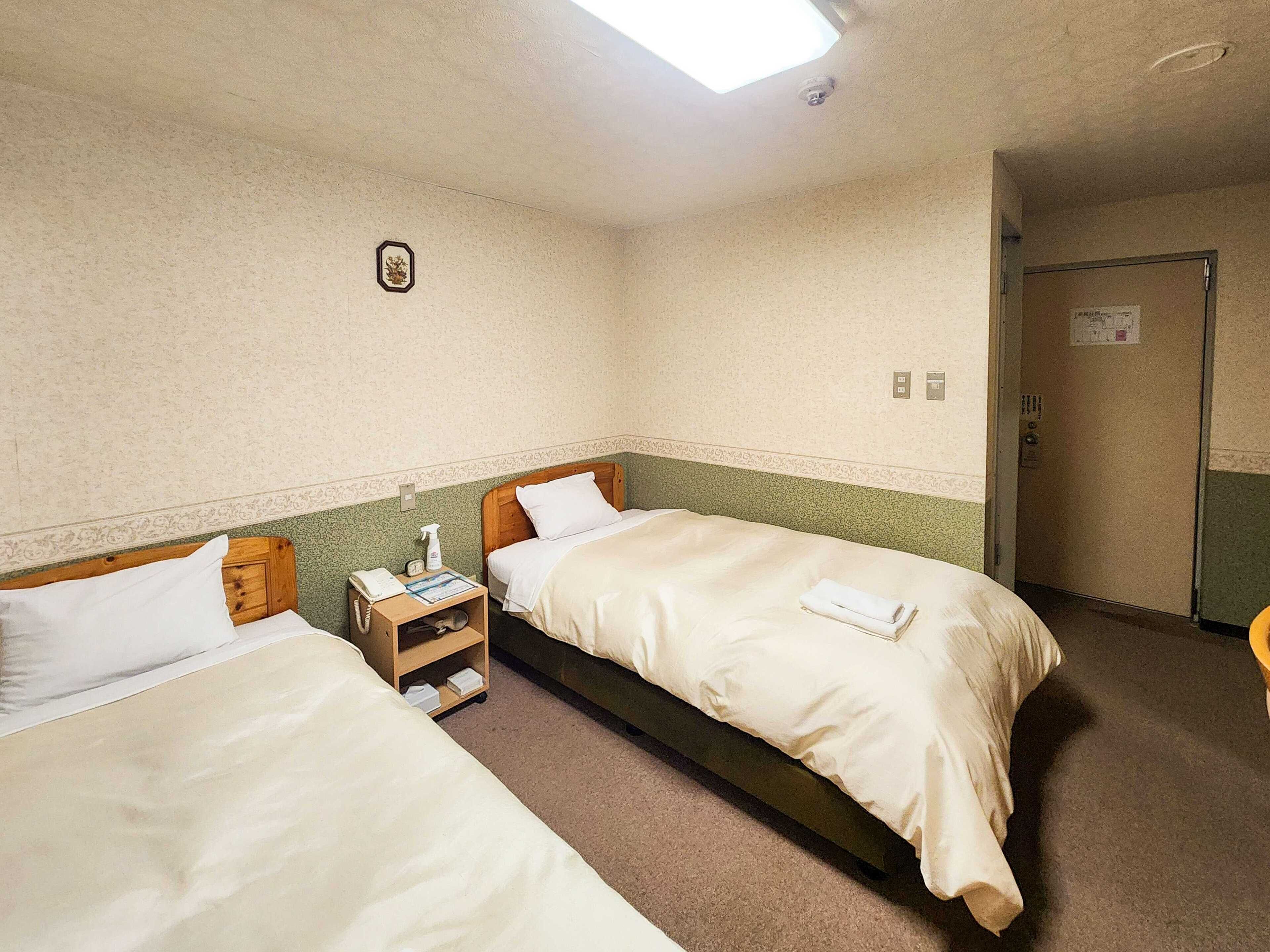 A hotel room with two single beds, a nightstand with a phone and a spray bottle, a small picture on the wall, and a door to the right.