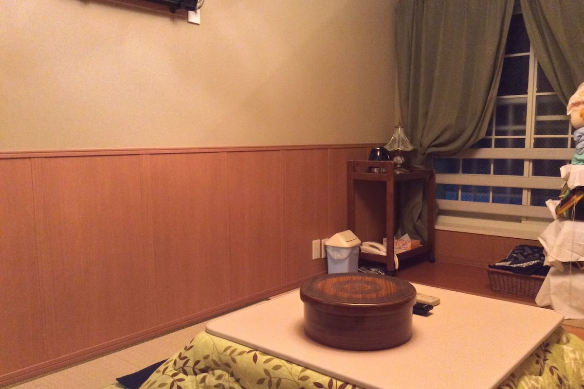 A room with a wooden floor, a small wooden side table with a lamp, a window with green curtains, and a bed with a patterned bedspread.