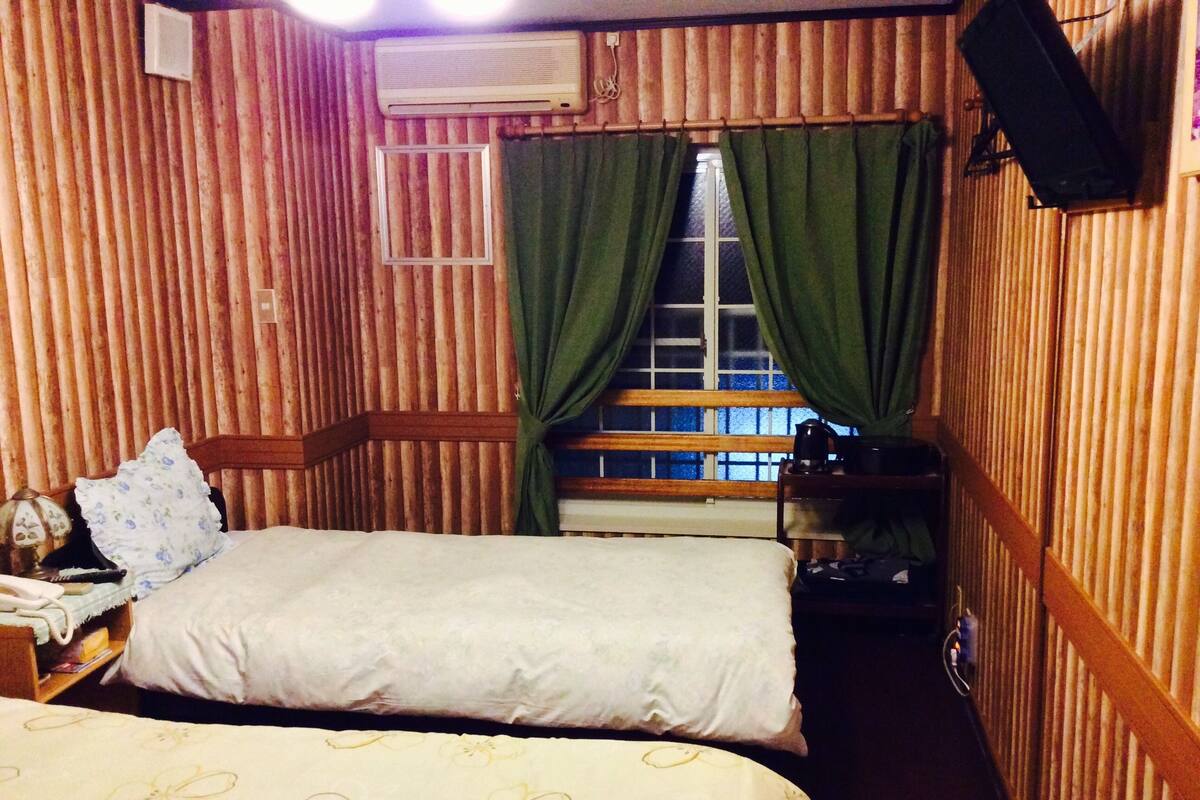 A room with two beds, a window with curtains, a radiator, and a wall-mounted speaker.