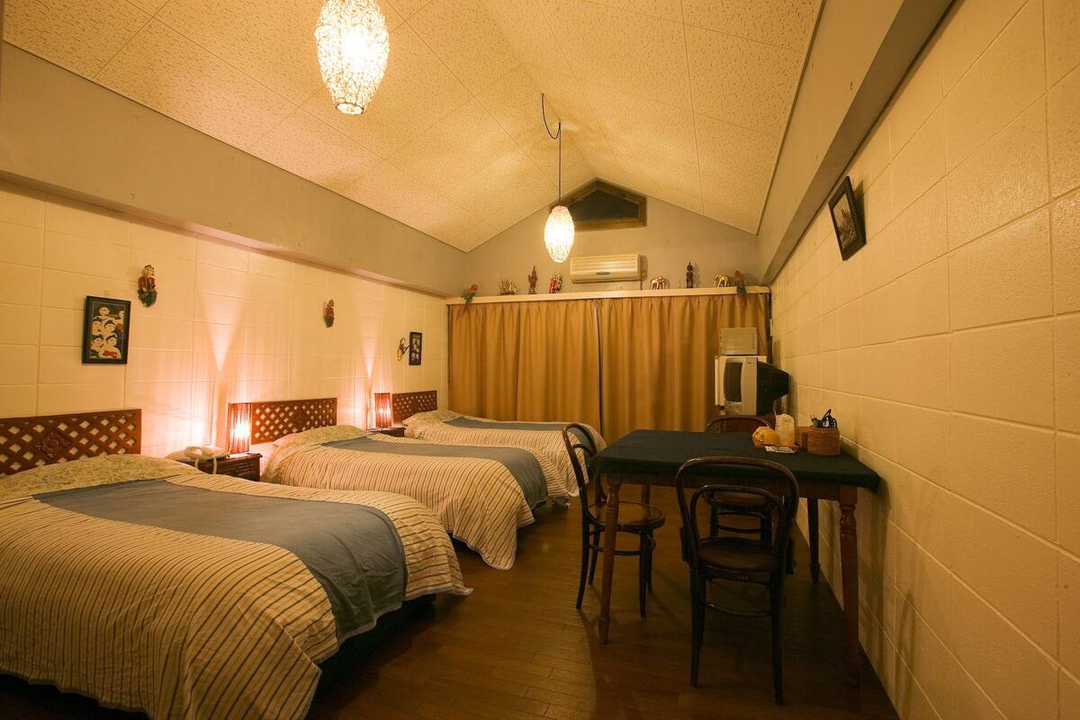 Quarto com duas camas, uma mesa, cadeiras e luminária no teto.