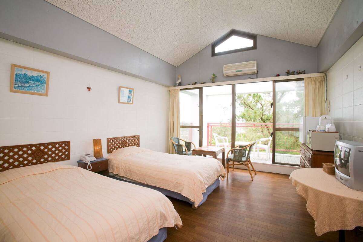 Quarto de hotel com duas camas, uma mesinha, televisão e varanda com vista para árvores.