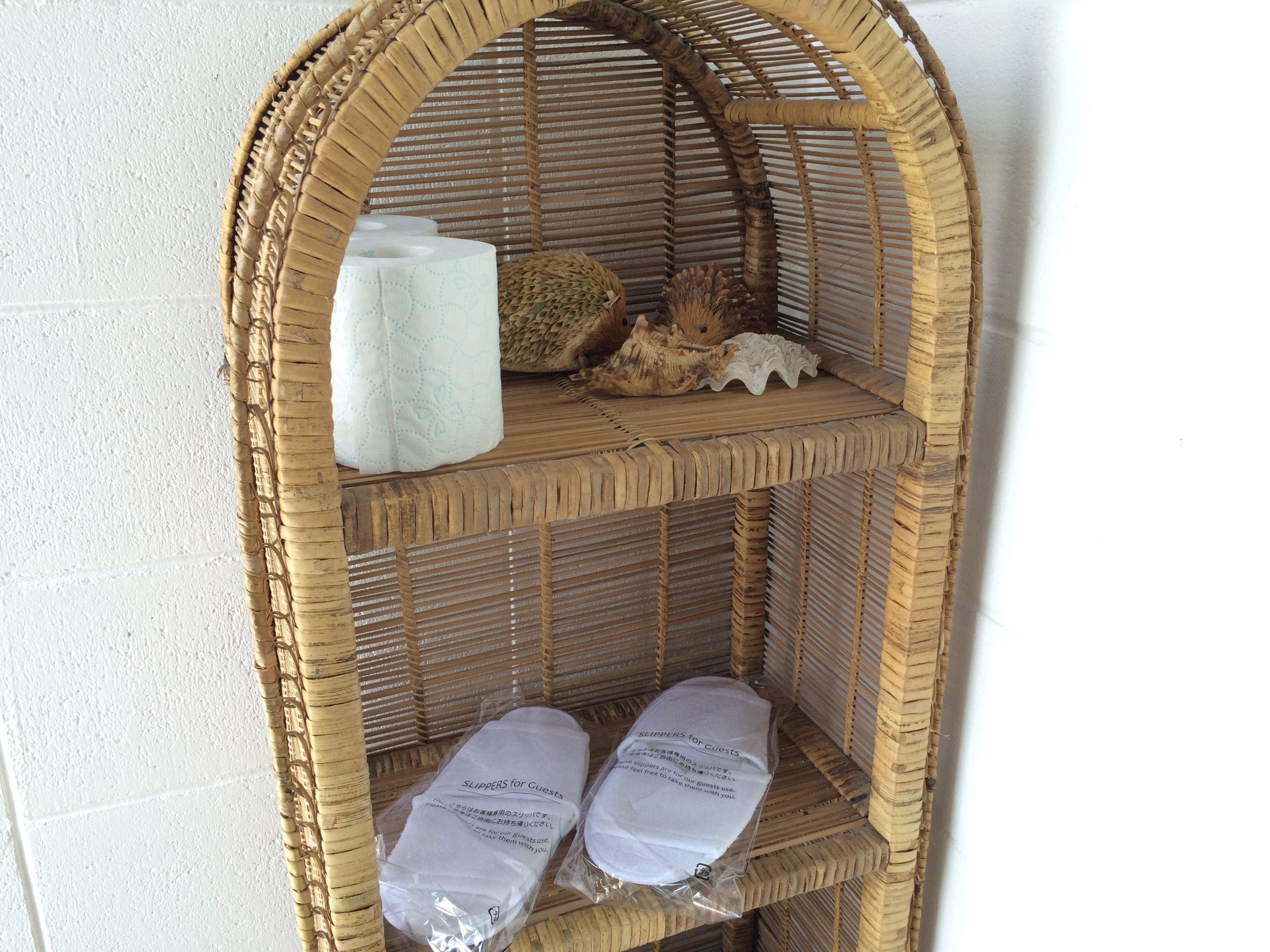 A wicker shelf with a rolled-up towel, a decorative shell, and two packages labeled "SLIPPERS for Guests".